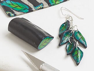 A sheet of polymer clay leaf cane rolled out, a stick of polymer clay leaf cane, and earrings featuring five drops of polymer clay leaves.