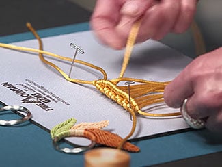 Hands working on a gold-colored macram&eacute; leaf design. A keyring with three macram&eacute; leaves is in the foreground.
